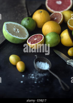 Preparing assorted citrus fruit for dessert with whole and halved grapefruit, oranges, limes, lemon and kumquat on a rustic kitchen counter with powdered sugar and a knife Stock Photo