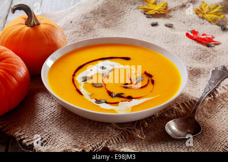 Delicious bowl of autumn or fall pumpkin soup garnished with cream and pumpkin pips on rustic burlap with two fresh pumpkins alongside Stock Photo