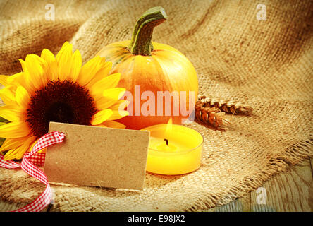Rustic warm toned Thanksgiving background with a blank card tied with a decorative red and white ribbon arranged with a fresh sunflower and pumpkin alongside a burning candle on rustic burlap fabric Stock Photo