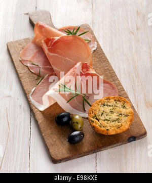 Italian prosciutto or parma ham in thin gourmet slices garnished with fresh rosemary and served with olives and golden toasted herb bread Stock Photo