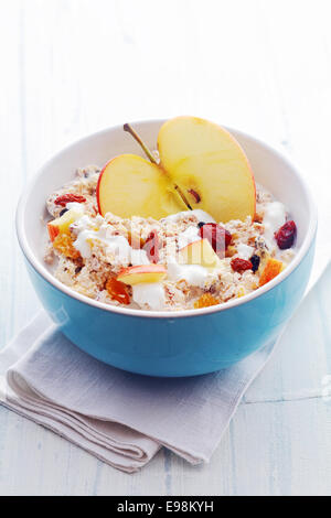 Bowl of healthy breakfast cereal with a halved fresh apple and topped with diced apple, dried fruit and nuts served with milk in a blue ceramic bowl, high angle view Stock Photo