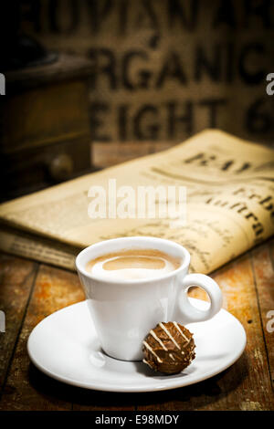 Cup of rich milky coffee with a chocolate bonbon on the saucer and a folded newspaper on the table behind in a coffee break concept Stock Photo