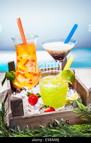 Trio of colourful tropical cocktails blended with fruit in different shaped glasses served overlooking a tropical beach Stock Photo
