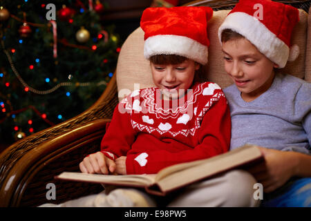 Adorable siblings in Santa caps reading book together on Christmas evening Stock Photo