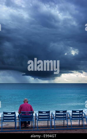 Elderly man sitting on bench at dyke and watching dark, menacing storm clouds over the sea Stock Photo