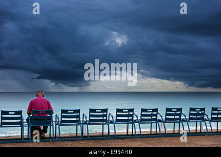 Elderly man sitting on bench at dyke and watching dark, menacing storm clouds over the sea Stock Photo