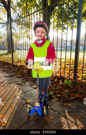 Child / girl on scooter in hi vis / high visibility vest jacket scootering / going to Reception class on a scooter at sunrise UK Stock Photo