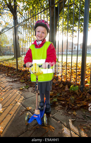 Child / girl on scooter in hi vis / high visibility vest jacket scootering / going to Reception class on a scooter at sunrise UK Stock Photo
