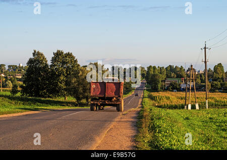 Autumn harvest field. Agricultural tractor with the trailer on the road near the village. Evening sunlight. Stock Photo