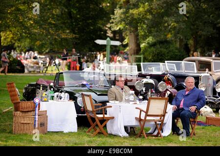 Two men sit at an elaborate picnic table set out  in front of an Aston Martin at a classic car concours Stock Photo