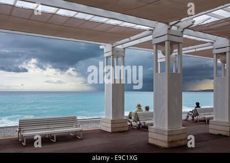 Elderly couple sitting on bench at promenade and watching dark, menacing rain clouds over the  Mediterranean sea at Nice, France Stock Photo