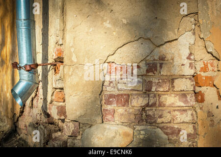 Cracked yellow wall of old building with visible red bricks. Few layers of materials. Metal hanging downspout. Shadows on wall. Stock Photo