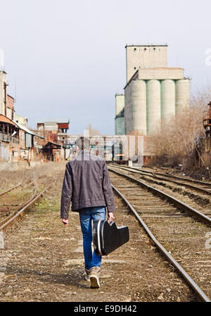 A young man with guitar case in hand going away by rails. Rear view Stock Photo