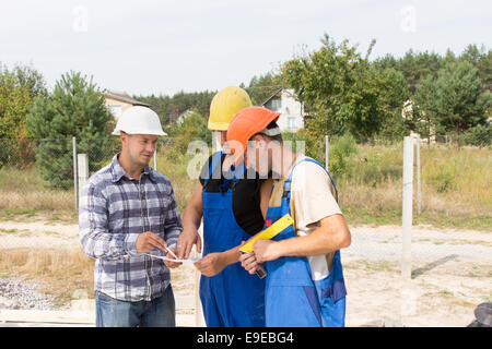 Engineer on a construction site discussing specifications with two workmen in overalls and hardhats as they group together aroun Stock Photo