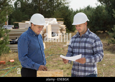 Two engineers having a discussion on site as they stand discussing a document with stacks of insulated wooden panels in view behind. Stock Photo