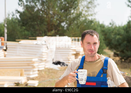 Workman taking a coffee break on a construction site standing in front of stacked building materials with a mug in his hand Stock Photo