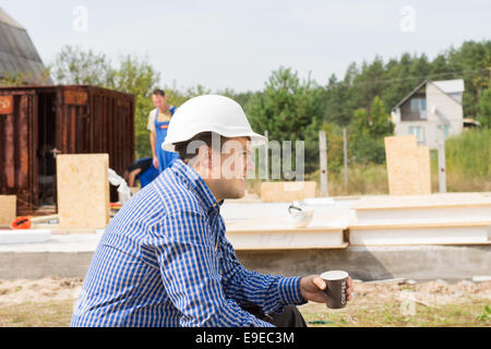 Thirsty builder taking a coffee break sitting side view with a mug of coffee in his hands as his crew continue work on the build Stock Photo