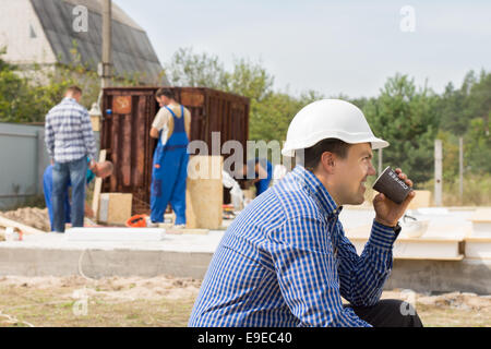 Workman sitting relaxing drinking coffee on a building site while his colleagues continue working, side view in a hardhat Stock Photo