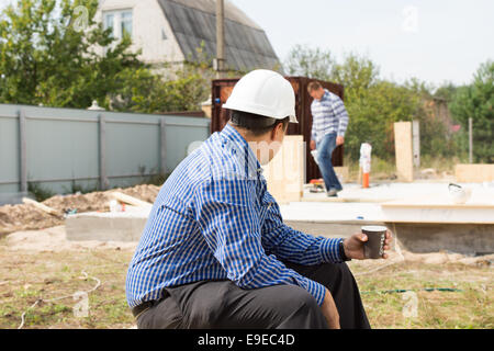 Builder on coffee break sitting on the construction site watching his colleagues continue working Stock Photo