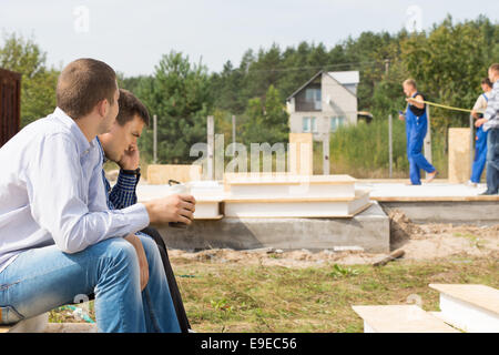 Middle Age Male Engineers Sitting on Corner at the Project Site with Construction Workers at the Background. Stock Photo