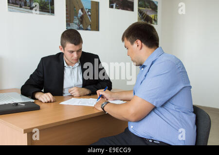 Male Client Signing Written Document on Wooden Desk of Young Real Estate Agent Inside the Firm. Stock Photo