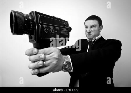 a black and white photo of a man holding a movie camera in a shooting a gun pose Stock Photo