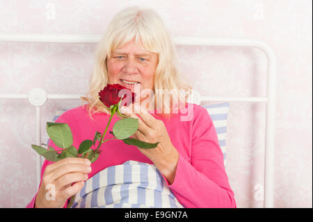 Cheerful woman in pink pajama sitting in bed with a red rose Stock Photo