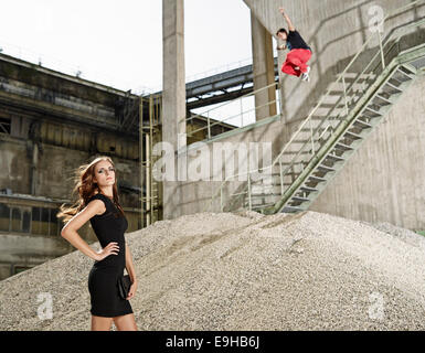 Young woman in black dress standing in front of a cement plant, young man jumping from a railing, Kiefersfelden, Bavaria Stock Photo