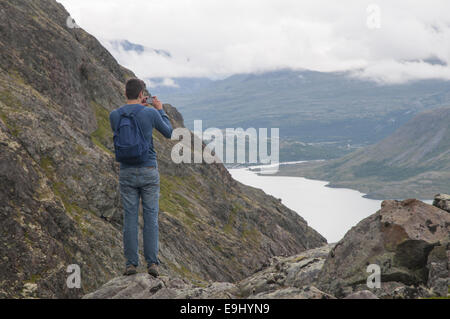 Young man taking a photograph on his phone standing on the edge of Besseggen mountain ridge overlooking Gjende lake and surrounding mountains. Norway. Stock Photo