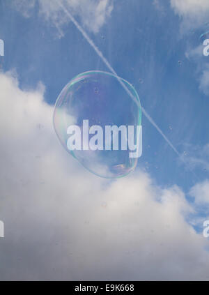 Giant bubble floating off into a blue cloudy sky with other smaller bubbles and jet vapour trail behind Stock Photo