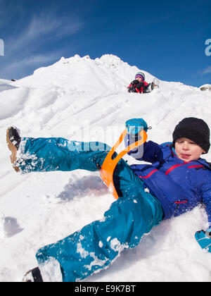 small  happy boy sledging fast down a steep snow covered hill with little girl and blue sky behind Stock Photo