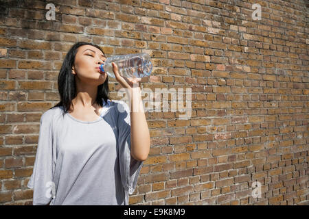 Beautiful young woman drinking water against brick wall Stock Photo