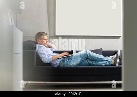 Full-length side view of Middle-aged man using laptop while lying on sofa Stock Photo