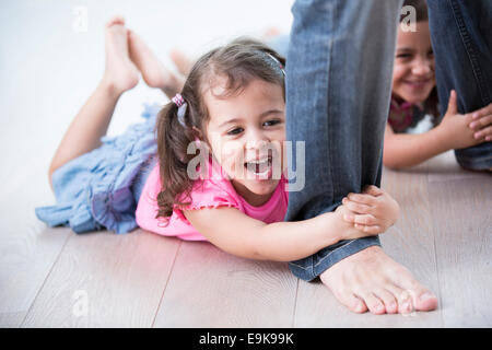 Playful girls holding father's legs on hardwood floor Stock Photo