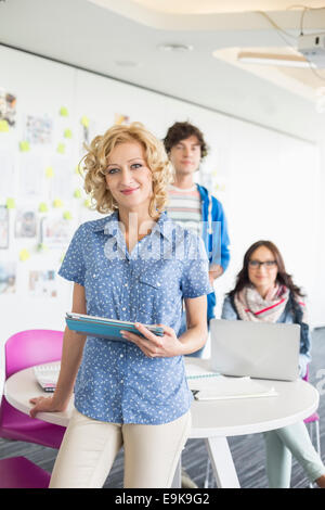 Portrait of beautiful businesswoman holding digital tablet with colleagues in background at creative office Stock Photo