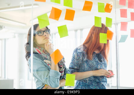 Creative businesswomen reading sticky notes on glass wall in office