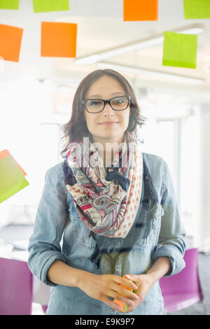 Portrait of smiling businesswoman standing by glass wall with sticky notes in office Stock Photo