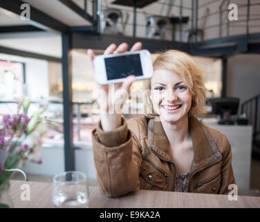Portrait of smiling young woman displaying cell phone in cafe Stock Photo