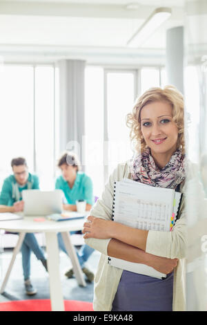Portrait of beautiful businesswoman holding files with colleagues working in background at creative office Stock Photo