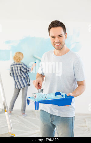 Portrait of man holding paint roller and tray with woman painting wall in background Stock Photo