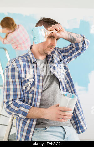Tired man holding paint can and brush with woman painting in background Stock Photo