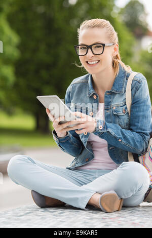 Full length portrait of smiling young female college student using tablet PC in park Stock Photo