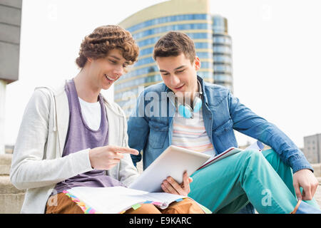 Happy young male college students using digital tablet against building Stock Photo