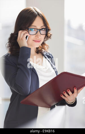 Portrait of smiling businesswoman using cell phone while holding file in office Stock Photo