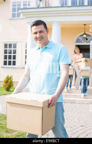Portrait of man carrying cardboard box while moving house with family in background Stock Photo