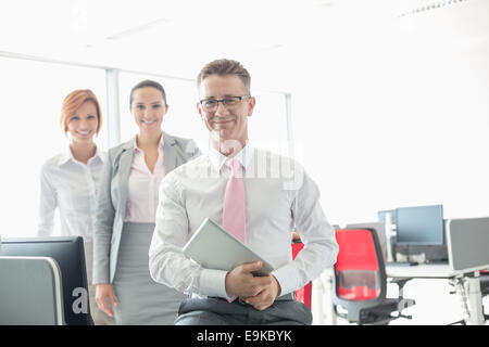 Portrait of happy businessman holding tablet PC with female colleagues in background at office Stock Photo