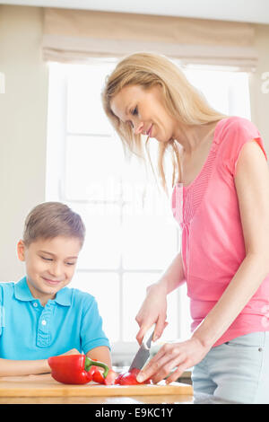 Boy looking at mother slicing red bell pepper in kitchen Stock Photo