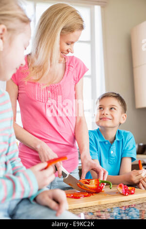 Happy woman chopping red bell pepper while standing with children in kitchen Stock Photo