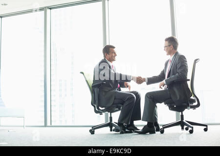 Full-length side view of businessmen shaking hands while sitting on office chairs by window Stock Photo