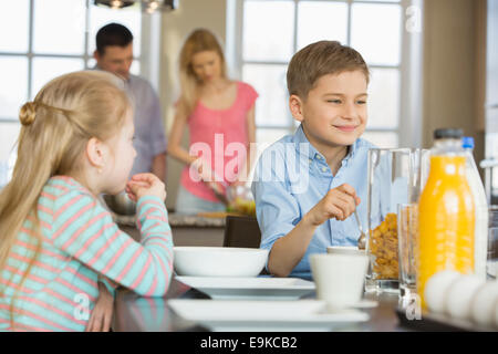 Siblings having breakfast at table with parents cooking in background Stock Photo